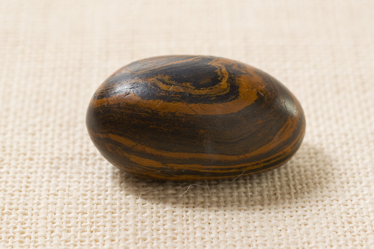 A smooth, oval-shaped stone with a polished surface featuring swirling patterns of dark brown and black, resting on a textured beige fabric background. The stone has a glossy finish that reflects light, highlighting its intricate natural striations.