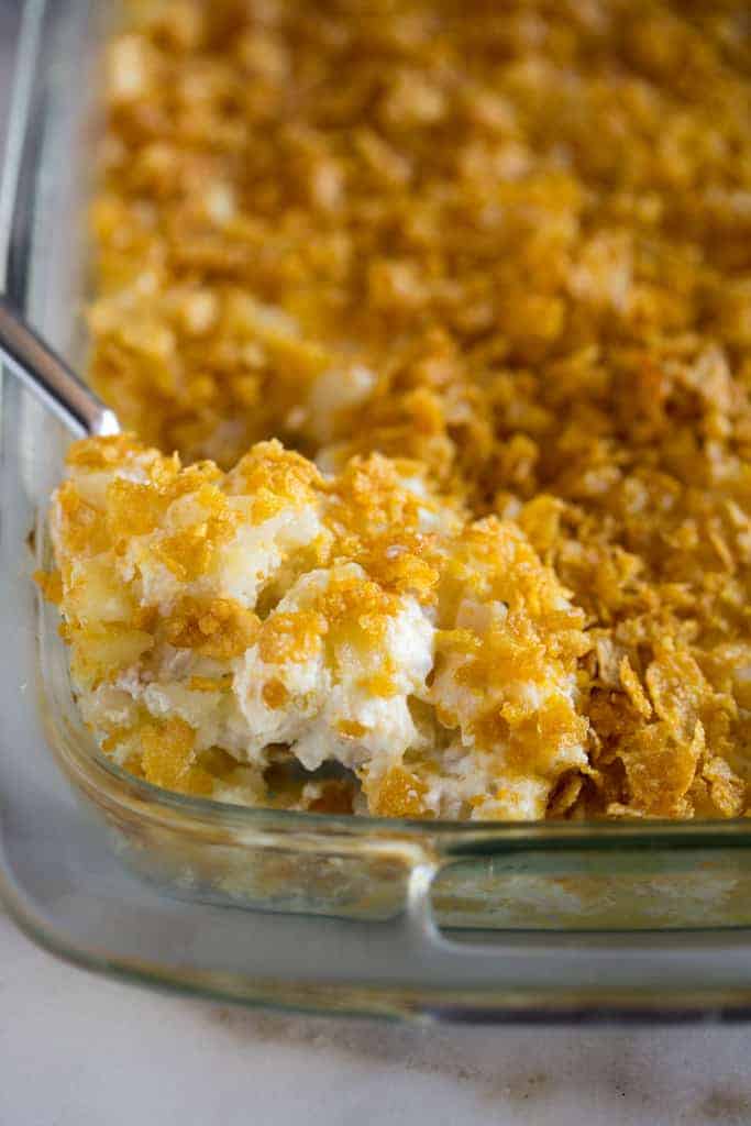 Close-up of a glass baking dish filled with Funeral Potatoes, a cheesy potato casserole topped with crushed cornflakes, with a serving spoon resting inside.