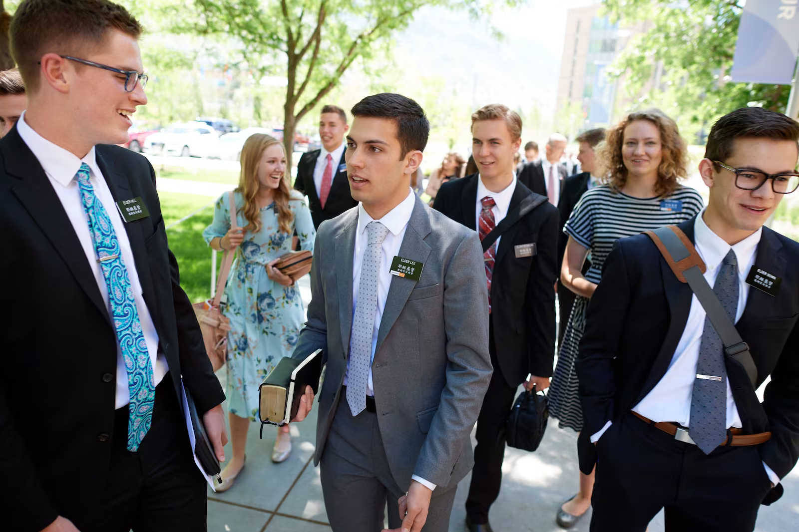 A group of young missionaries at the Provo Missionary Training Center walking outdoors on a sunny day. The men are dressed in suits and ties, and the women are wearing dresses. Many of the missionaries wear name tags and carry scriptures or notebooks as they walk along a tree-lined pathway with buildings and mountains visible in the background.