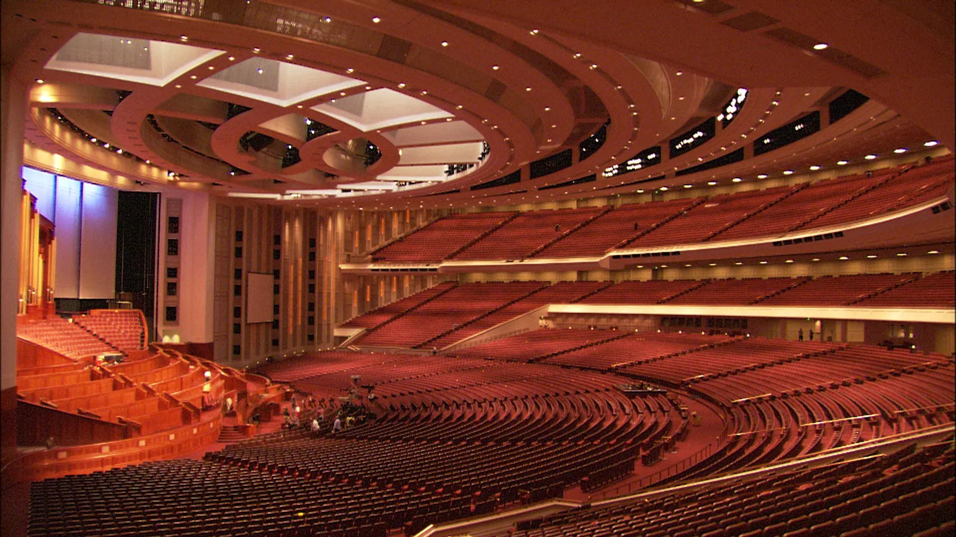 Interior of the Conference Center in Salt Lake City. The vast auditorium features curved rows of red seating, tiered balconies, and warm wood paneling around the rostrum. Completed in 2000, the building can seat over 21,000 people for General Conference and other Church events.
