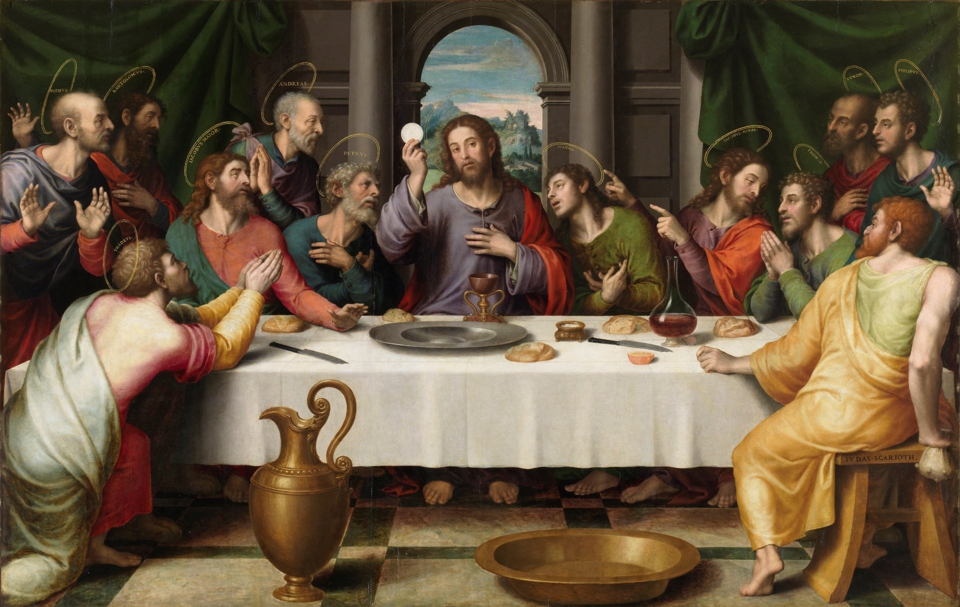 A painting by Juan de Juanes titled The Last Supper shows Jesus at the center of a long table, holding up the Eucharistic bread as He institutes the sacrament. The apostles surround Him, reacting with gestures of surprise, devotion, and conversation. Each apostle is identified by a golden halo with their name. Judas Iscariot, seated at the front right without a halo, clutches a money bag and looks away. The scene is richly colored, with bread, wine, and ceremonial vessels arranged on the white-clothed table, framed by green drapery and a view of a landscape through an arched window.