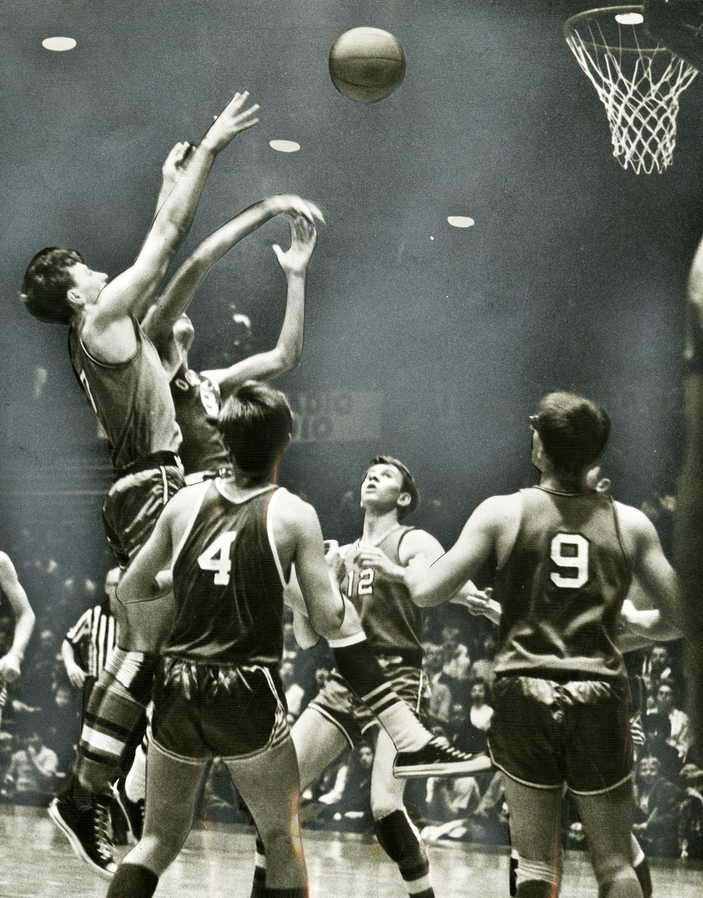 Black-and-white action shot of young men playing in the All-Church Basketball Tournament. Players in numbered jerseys jump and reach for a rebound beneath the hoop, while a crowd watches from the stands. The Highland and Downey 4th Ward teams are competing for the Junior Title.