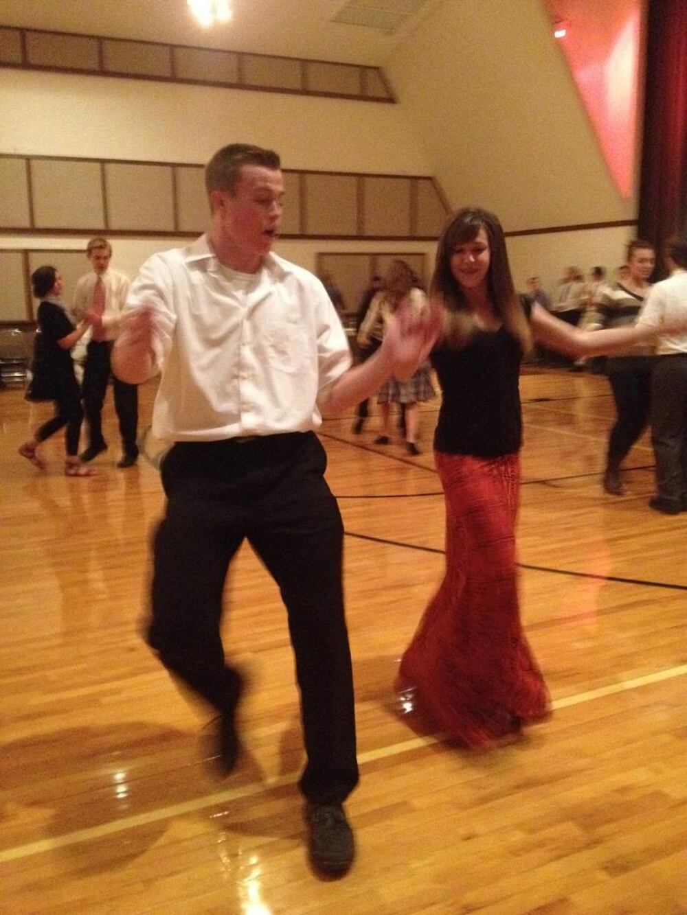 A candid photo of a young man and woman dancing enthusiastically in a gymnasium with polished wooden floors. The man, dressed in a white shirt and dark pants, appears mid-step with an expressive face and raised arms. The woman, wearing a black top and a long red patterned skirt, smiles as she dances beside him. Other couples can be seen dancing in the background. The setting has high ceilings and acoustic panels along the walls.