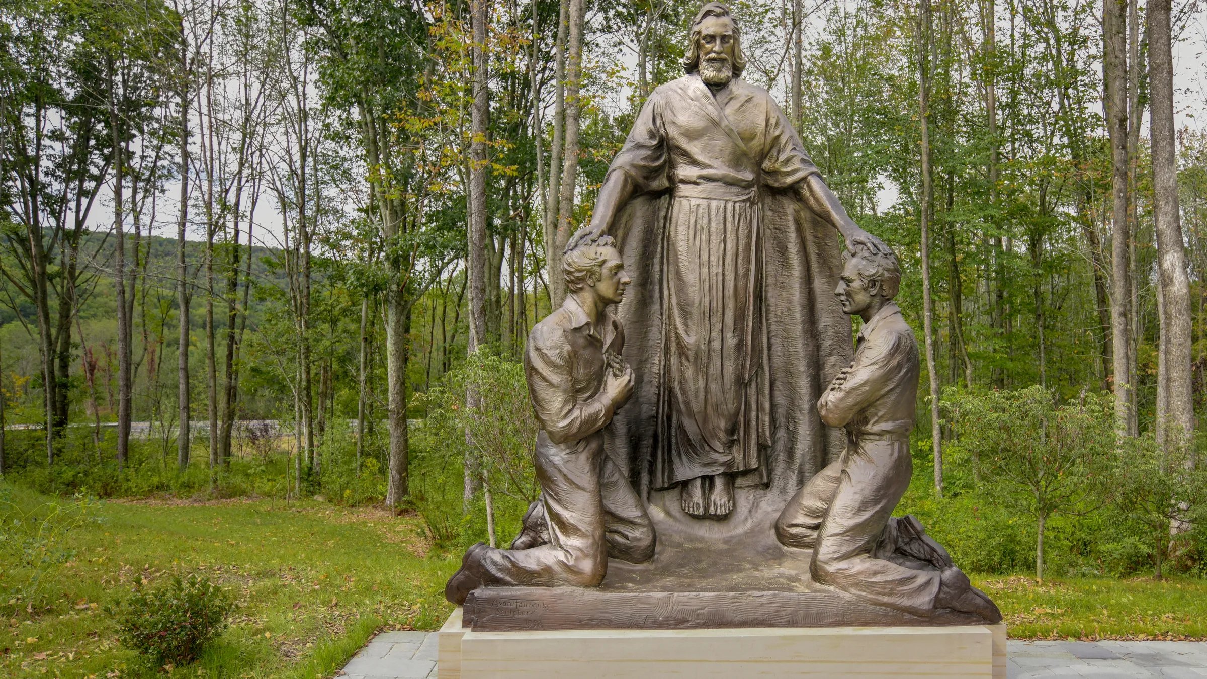 Bronze statue in a forest clearing showing a bearded man standing with his arms extended, placing his hands on the heads of two kneeling men. The figures are set on a stone base, surrounded by grass, trees, and a distant view of hills.