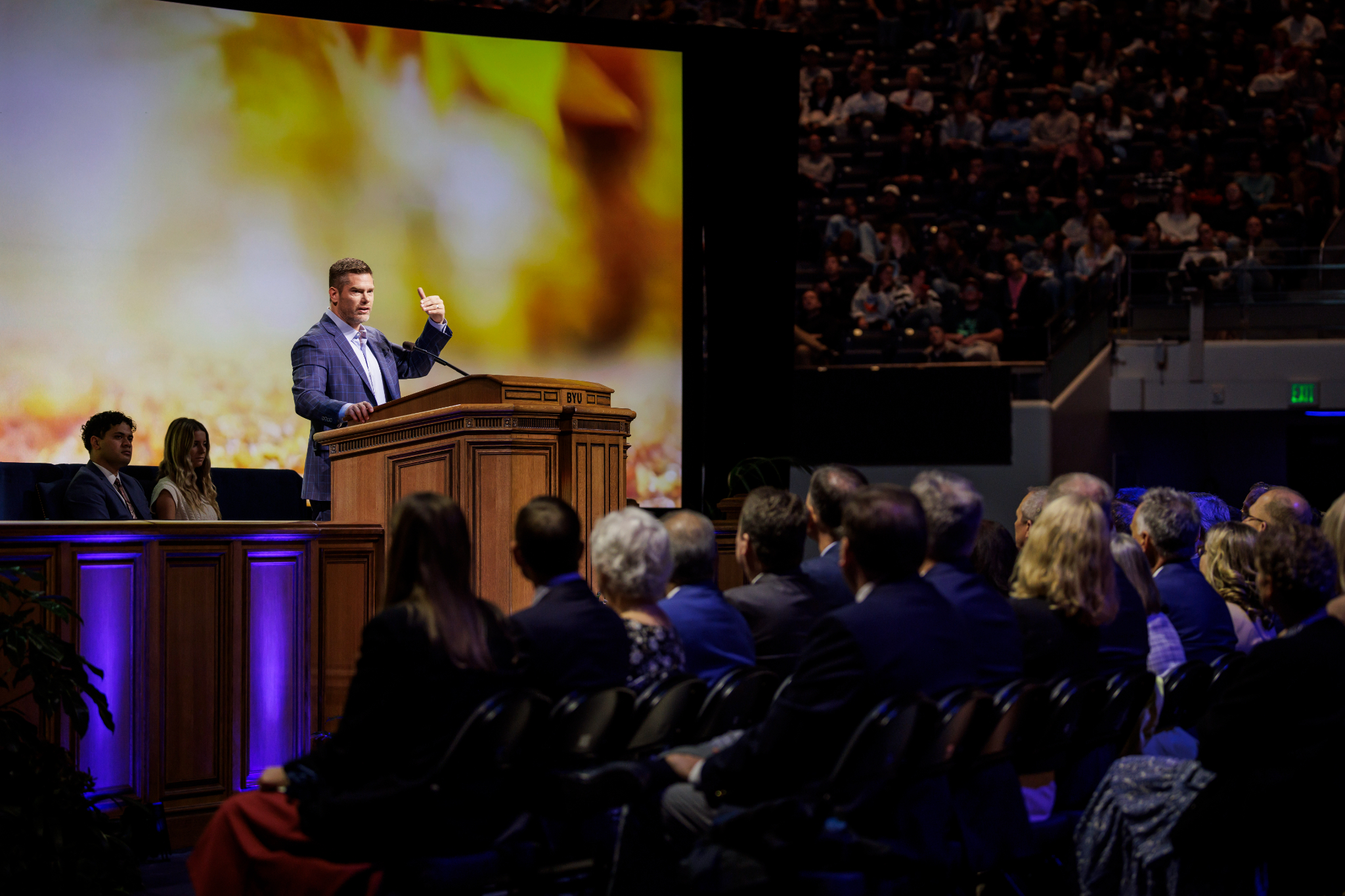 A man in a plaid suit speaks passionately at a wooden podium labeled 'BYU' in front of a large audience. A bright, blurred background image is displayed on a giant screen behind him. People are seated on stage behind the speaker, while a large audience listens in the dimly lit auditorium.