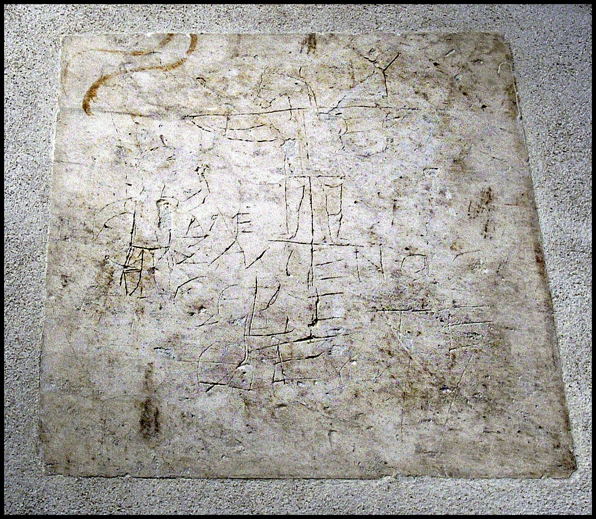 Ancient scratched graffiti etched into a rectangular slab of stone. The carving depicts a crude human figure with a donkey's head, arms outstretched on a cross, accompanied by a smaller figure standing to the left, raising one hand. Greek letters are scrawled beneath the figures, forming an inscription. The surface of the stone is weathered and uneven, with faint markings and discoloration throughout.