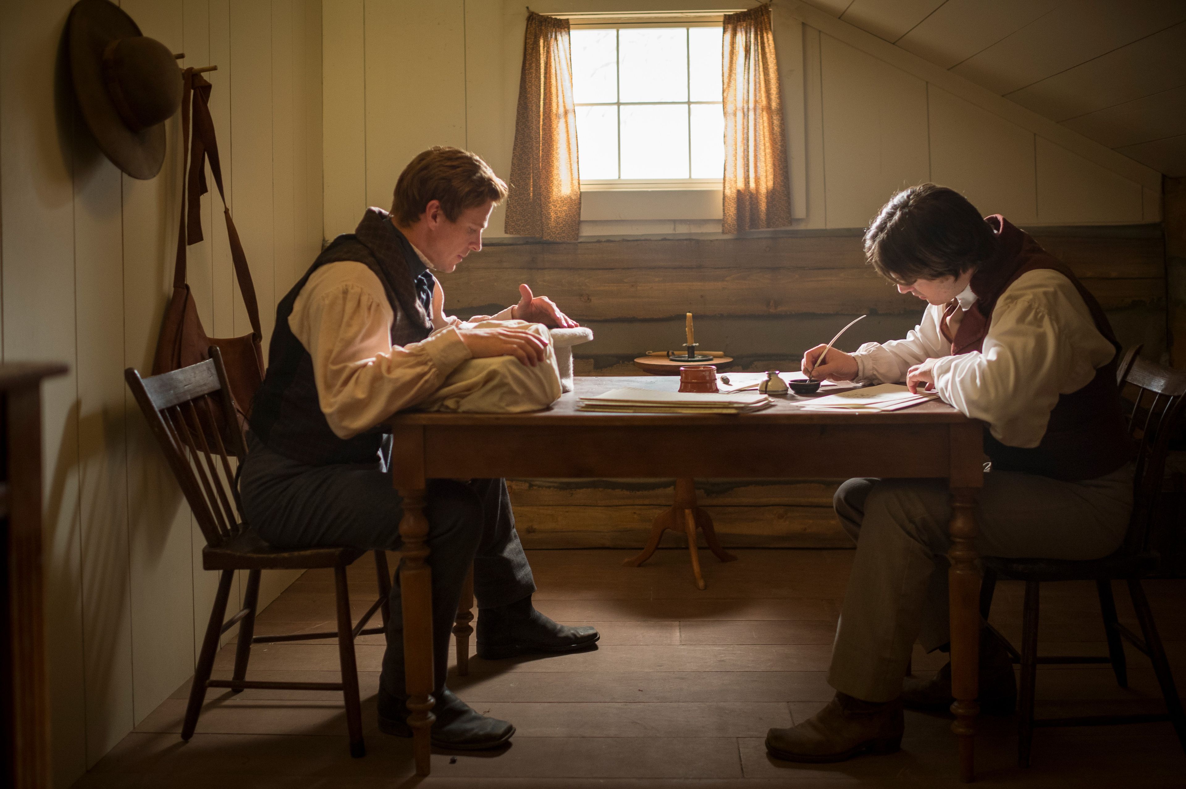 Two men in nineteenth-century attire sit at a wooden table in a dimly lit room. One examines an object in a hat next to the golden plates wrapped in cloth, while the other writes with a quill.
