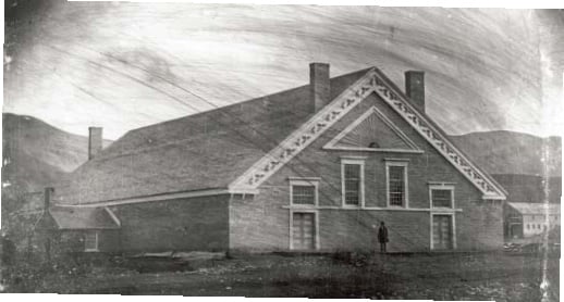 Photograph of the Old Tabernacle, 1850s. Sometimes called the Adobe Tabernacle, this early meetinghouse stood on the south end of Temple Square in Salt Lake City. A single figure stands in front of the large brick structure with decorative gable trim, surrounded by mountains in the background.