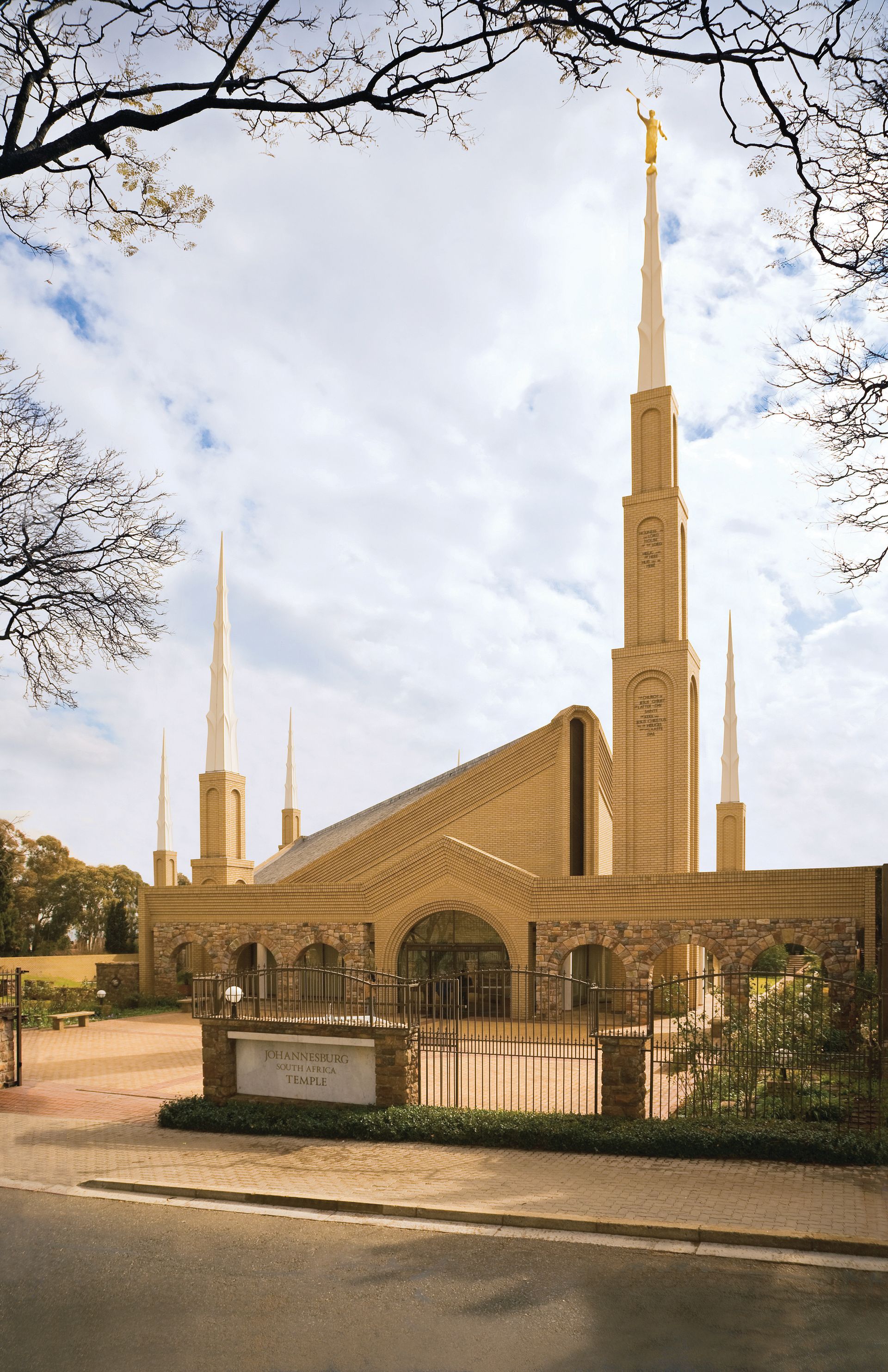 Exterior view of the Johannesburg South Africa Temple of The Church of Jesus Christ of Latter-day Saints, featuring its distinctive golden spire topped with the Angel Moroni statue. The temple is constructed with light-colored brick and stone, and surrounded by a gated entrance and landscaped grounds. Leafless tree branches frame the sky above.