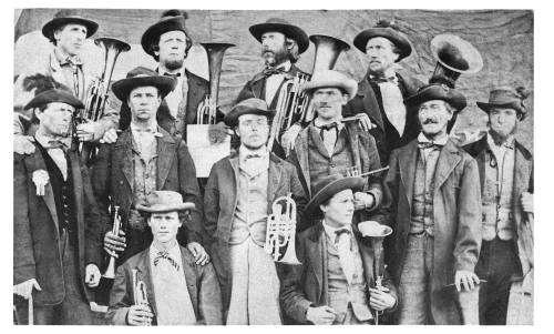 A black-and-white historical photograph of a 19th-century brass band. Thirteen men are dressed in period clothing, including coats, vests, cravats, and wide-brimmed hats. They pose with their instruments, including cornets, trombones, euphoniums, and tubas. Some of the men have serious expressions, while others smile slightly. The background appears to be a plain cloth or tarp, drawing focus to the musicians.