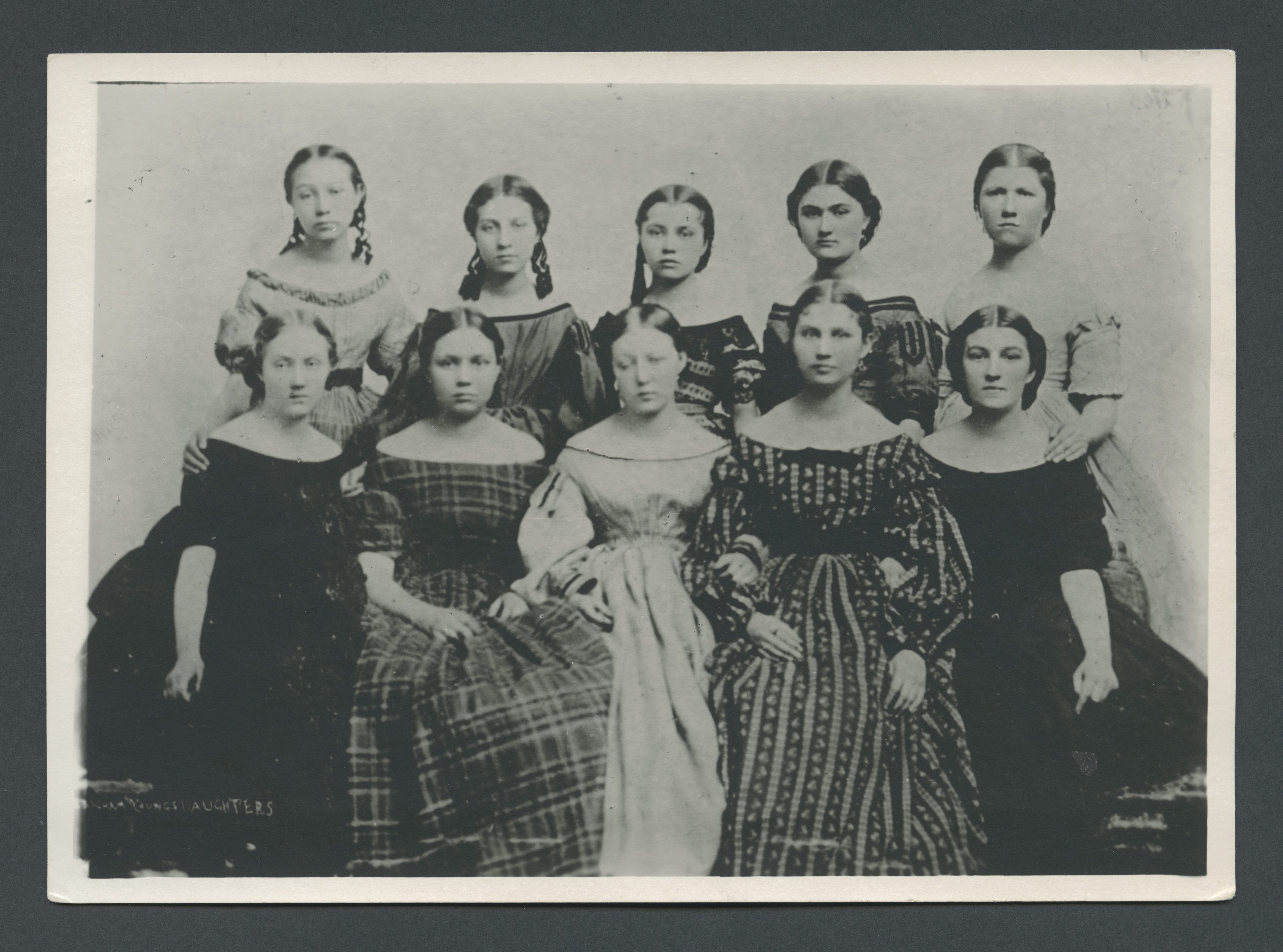 A vintage black-and-white photograph of nine young women posing formally in two rows, with four seated in the front and five standing in the back. They are dressed in 19th-century gowns with various patterns, including plaid and intricate designs, and have their hair neatly styled. The women have serious expressions and are looking directly at the camera. The bottom left corner of the image contains faint text that reads "Brigham Young's Daughters." The background is plain.