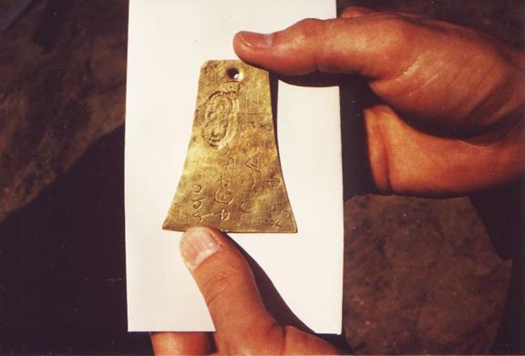 A photograph of two hands holding the last known Kinderhook plate, a small bell shaped piece of brass that was a forgery of an ancient artifact