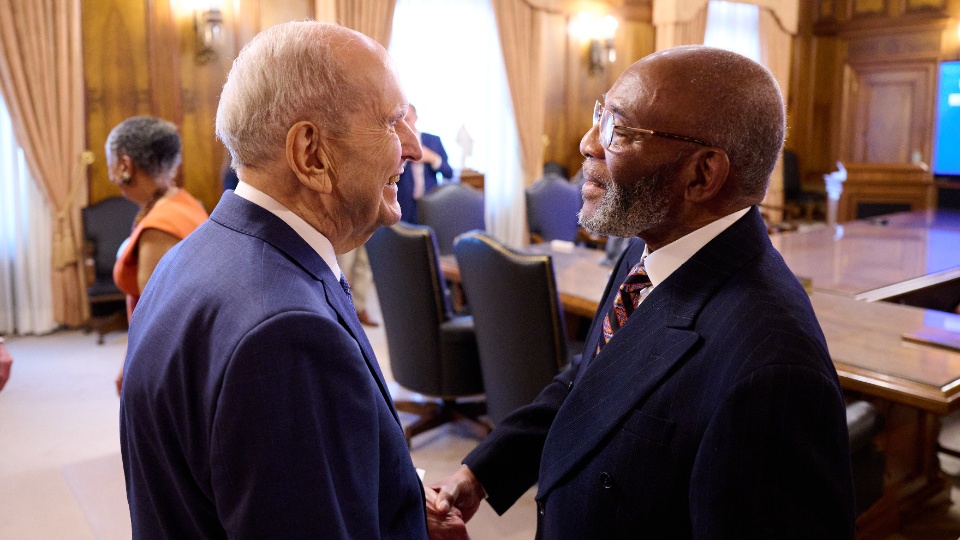 Two elderly men in suits shake hands and smile at each other in a formal meeting room. A woman stands in the background near a conference table with high-backed chairs and curtained windows.