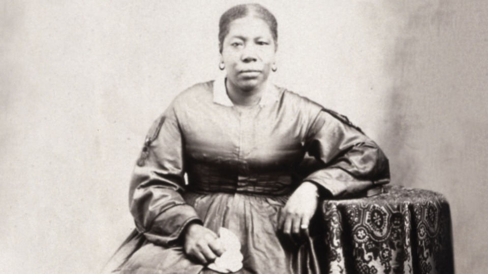 A historical black-and-white portrait of a Jane Manning James sitting. She wears a full-skirted satin dress with long sleeves, a fitted bodice, and a white collar. Her hair is neatly pulled back, and she wears simple hoop earrings. One elbow rests on an ornately patterned tablecloth, while the other holds a white handkerchief in her lap. The background is plain.