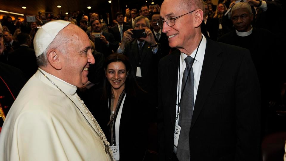 Pope Francis, wearing white robes, smiles and converses with President Eyring, wearing a dark suit and tie. Both are wearing conference badges in a crowded auditorium filled with people. Attendees in the background are taking photos and watching the interaction.