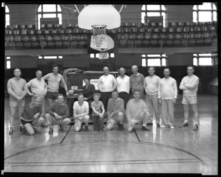 Black-and-white photo of the Deseret Gym Basketball Team posing on a gym floor beneath a basketball hoop, taken on December 22, 1948. Sixteen men and two boys stand and kneel in front of a cart decorated with a sign that reads "Merry Xmas to You Carroll Bob." The photo captures a holiday moment in a vintage basketball court with wooden bleachers in the background.