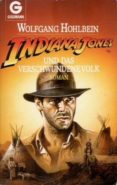 A book cover of Indiana Jones und das verschwundene Volk by Wolfgang Hohlbein. The cover features a painted illustration of Indiana Jones wearing his iconic fedora and brown leather jacket, with a serious and determined expression. The background depicts a desert-like setting with teepees, Native American figures, and a warm, orange-toned sky. The title is displayed in bold, stylized lettering with "Indiana Jones" in yellow and red, and the author's name in white at the top. The publisher's logo, "Goldmann," is in the upper left corner.