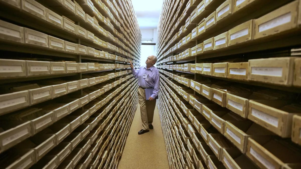 A man, Gary Mokotoff, a Jewish geneaologist, looks at name registers in the middle of a lot of shelves, in Germany, 2008.