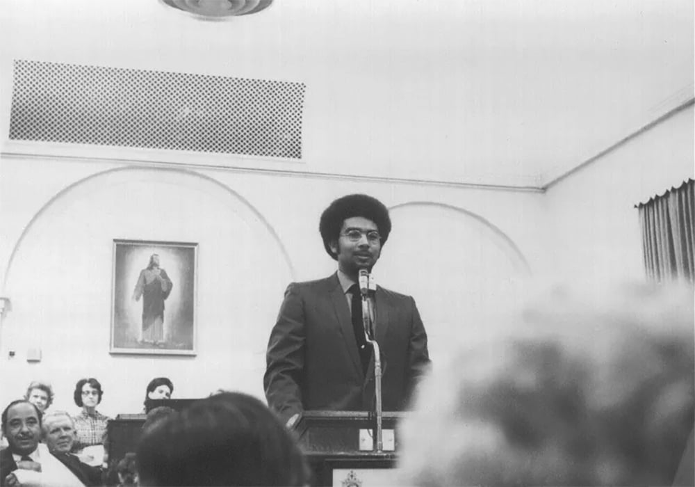 In a black and white photograph, Darius Gray, original counselor in the presidency of the Latter-day Saint Genesis Group, an organization for African American members of the Church, speaks at their first meeting in 1971, with a picture of Jesus Christ on the wall behind him.