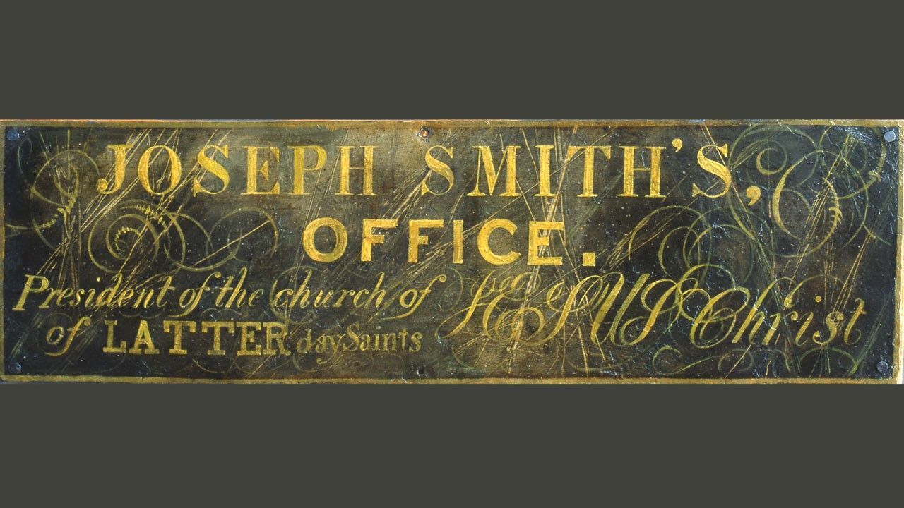 Historic painted sign in Joseph Smith's Red Brick Store reading 'Joseph Smith's Office. President of the church of Jesus Christ of LATTER day Saints' in gold lettering on a dark background, with ornate script flourishes and decorative scrollwork.