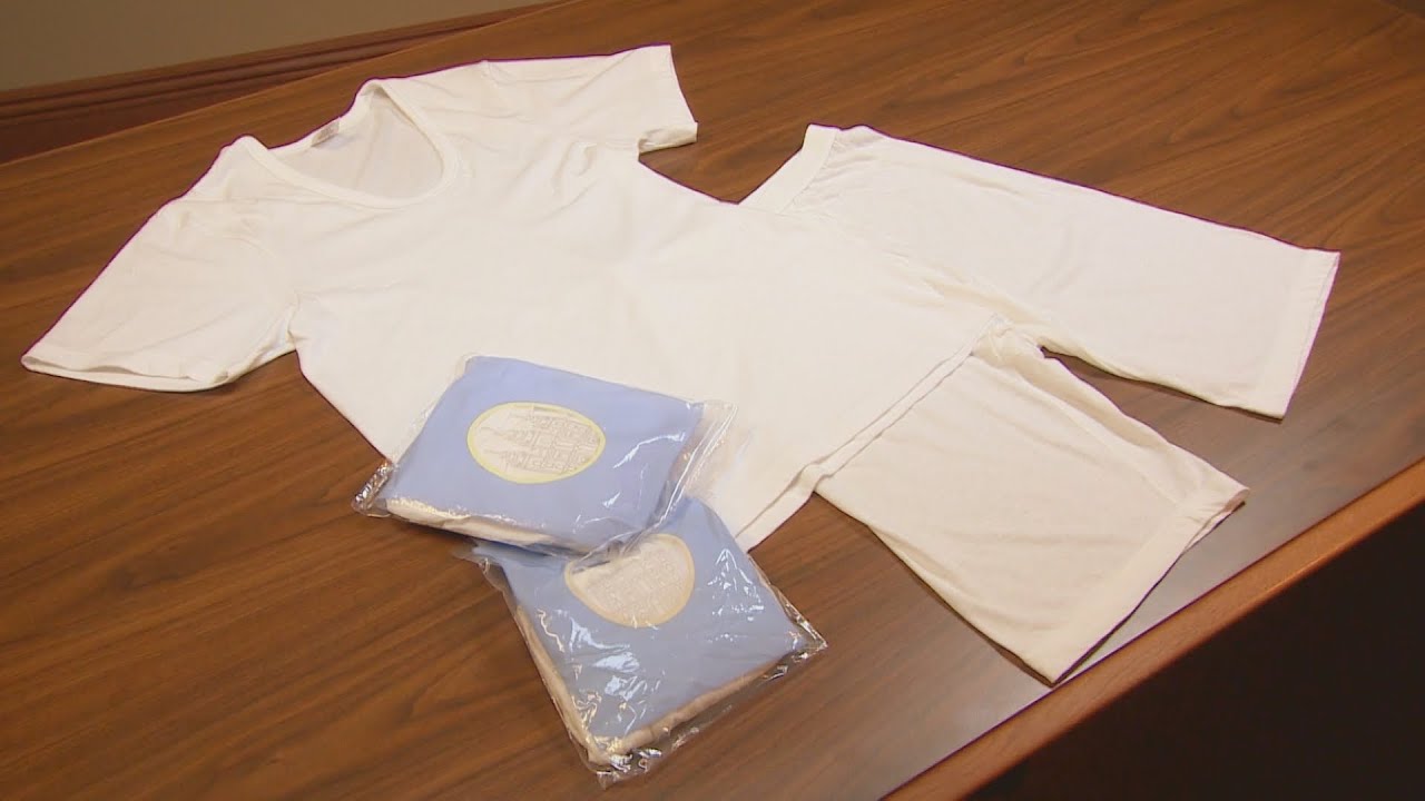 A set of white temple garments, consisting of a short-sleeved top and knee-length bottom, laid out on a wooden table. Two additional packaged sets of garments, wrapped in blue plastic with a gold emblem, are placed beside them.