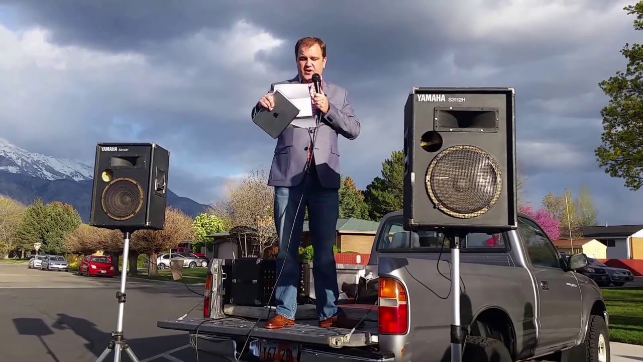 A color photograph from April 26, 2016, of Jeremy Runnells, author of the CES Letter, announcing that he is leaving the Church of Jesus Christ of Latter-day Saints, on top of a pickup truck tailgate, PA speakers on both sides, in church parking lot, mountains in background.
