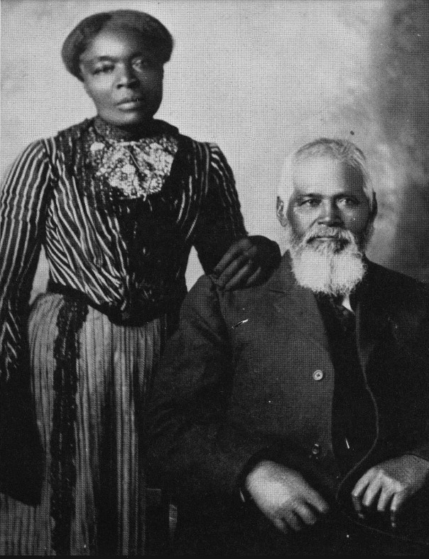 A historical black-and-white photograph of Samuel and Amanda Chambers. Samuel, an older man with a full white beard and short white hair, is seated wearing a dark suit with a unbuttoned coat. Amanda stands beside him with her hand resting gently on his shoulder. She wears a striped blouse with lace detailing at the collar and a long, pleated skirt.