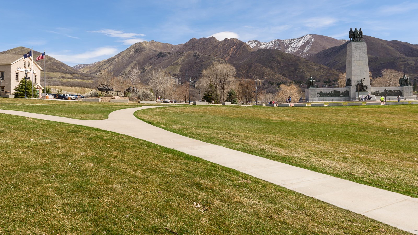 Wide view of the "This Is the Place" Monument in Heritage Park, Salt Lake City, Utah. The monument, featuring bronze pioneer figures atop a large stone pedestal, stands against a backdrop of the Wasatch Mountains. A curved sidewalk leads from the visitor center on the left toward the monument on the right, with grassy fields and early spring trees in the foreground.