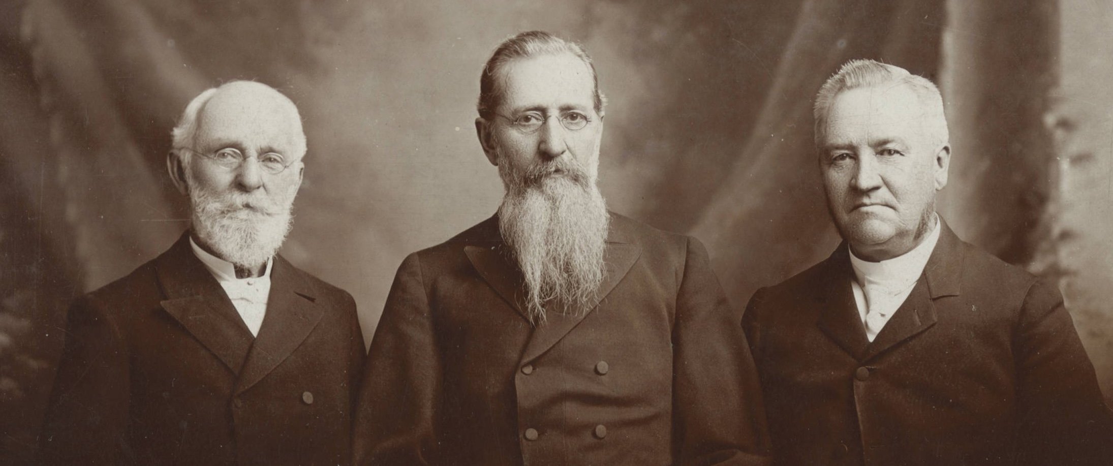 A sepia-toned historical photograph of three elderly men in formal 19th-century attire, standing side by side. All three have facial hair: the man on the left has a white beard and glasses, the man in the center has a long, full white beard and glasses, and the man on the right has short white hair and no beard. The background is a simple studio backdrop with soft lighting and a neutral tone.