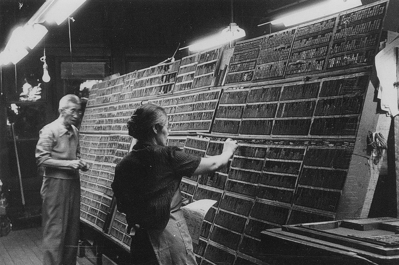 A black-and-white photograph of two people working in a traditional print shop. They are standing in front of a massive wall filled with rows of small compartments containing metal movable type, used for letterpress printing. The woman in the foreground selects type from the compartments, holding a paper in one hand. The man in the background appears to be organizing or preparing type as well. Overhead fluorescent lights illuminate the workspace, casting shadows on the walls and floor.