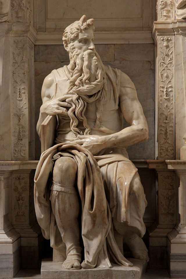 A marble statue of Moses, sculpted by Michelangelo, seated with a long, flowing beard and muscular arms. The figure has two horns on his head, a common artistic interpretation due to a translation of biblical texts. Moses holds part of his beard in his right hand while his left arm rests on the stone surface. The statue is set within an ornate architectural niche.