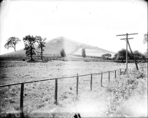 Eine Schwarz-Weiß-Fotografie des Hügels Cumorah in New York aus dem Jahr 1907, die einen grasbewachsenen Hang mit einem von Bäumen gesäumten Weg, eine eingezäunte Weide im Vordergrund und hölzerne Strommasten an der Seite zeigt.