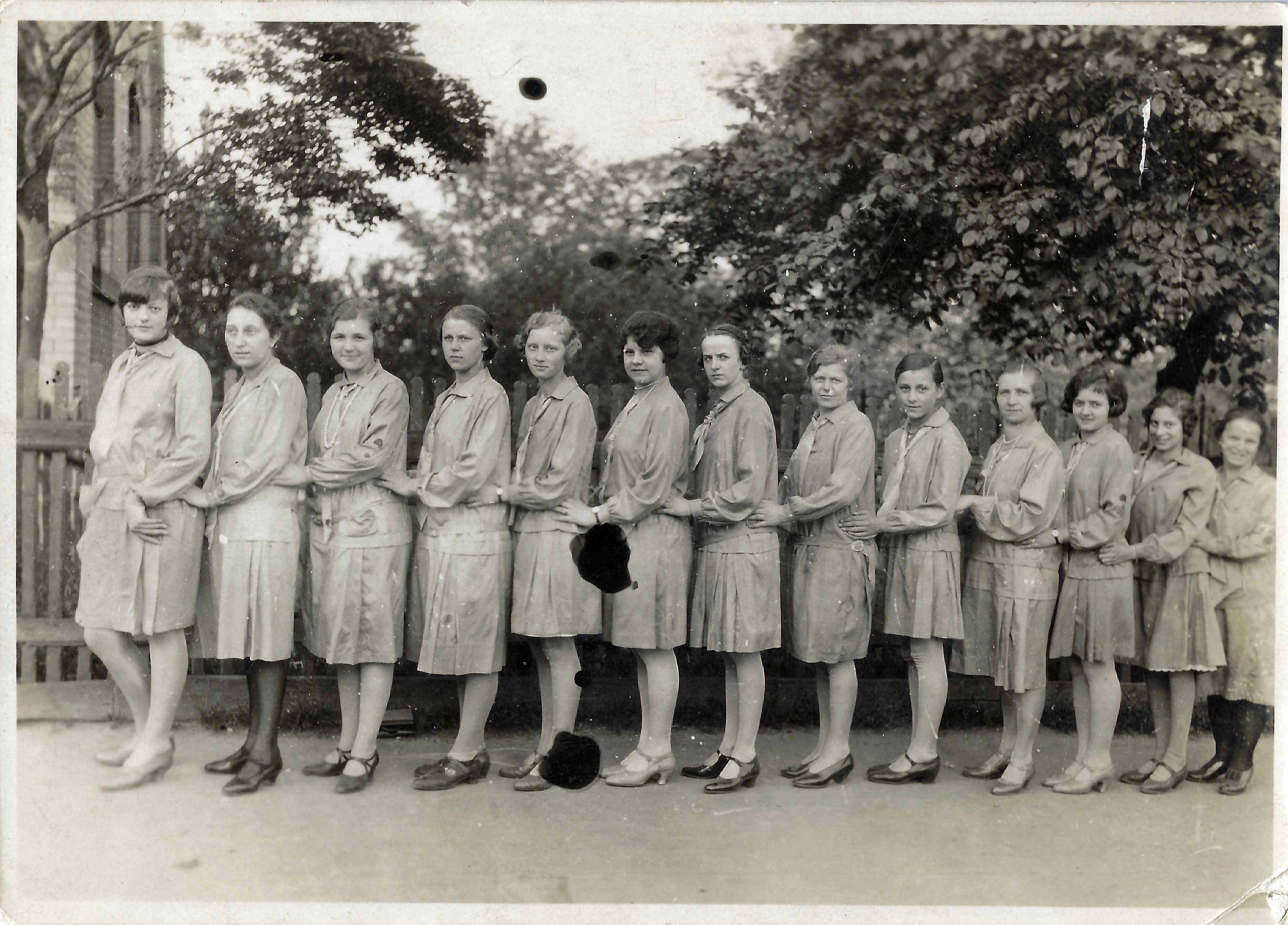 A vintage black-and-white photograph of a group of thirteen young women standing outdoors in a line, each placing their hands on the waist or back of the person in front of them. They are dressed in matching knee-length dresses with long sleeves, likely from the early 20th century, paired with stockings and various styles of heeled shoes. The background features trees, a wooden fence, and part of a brick building. Some of the women are smiling, while others have more serious expressions. The photograph shows signs of age, with visible stains and spots.